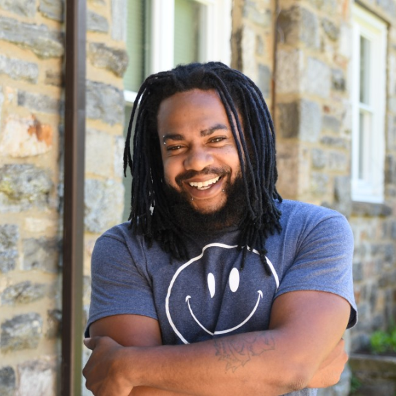 A dark toned man with dreadlocks wearing a blue t-shirt with a white smiley face stands on a porch